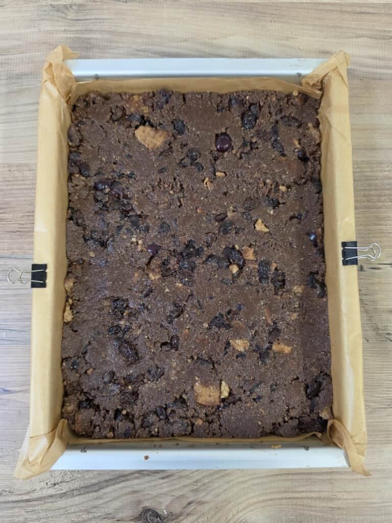A rectangular tin lined with greaseproof paper filled with raw, unbaked chocolate dough containing scattered dried fruit and biscuit pieces.