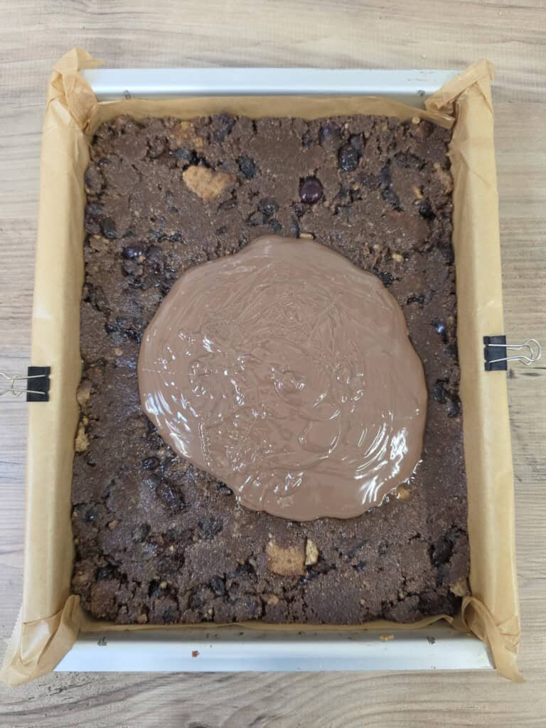 A square baking tin lined with greaseproof paper containing a dark, textured chocolate dessert mixture with a pool of melted chocolate in the centre.
