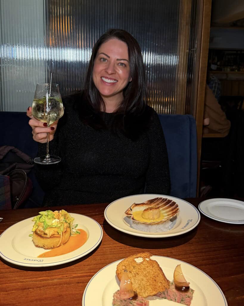 A smiling woman holds up a glass of white wine at a restaurant table set with three plates of food in this Laurenscravings scene.