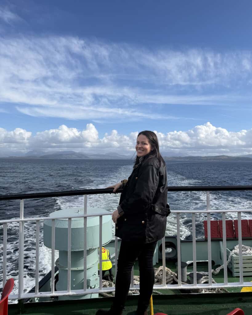 A woman smiles whilst standing at the railing of a ferry boat looking out over the ocean towards distant mountains.