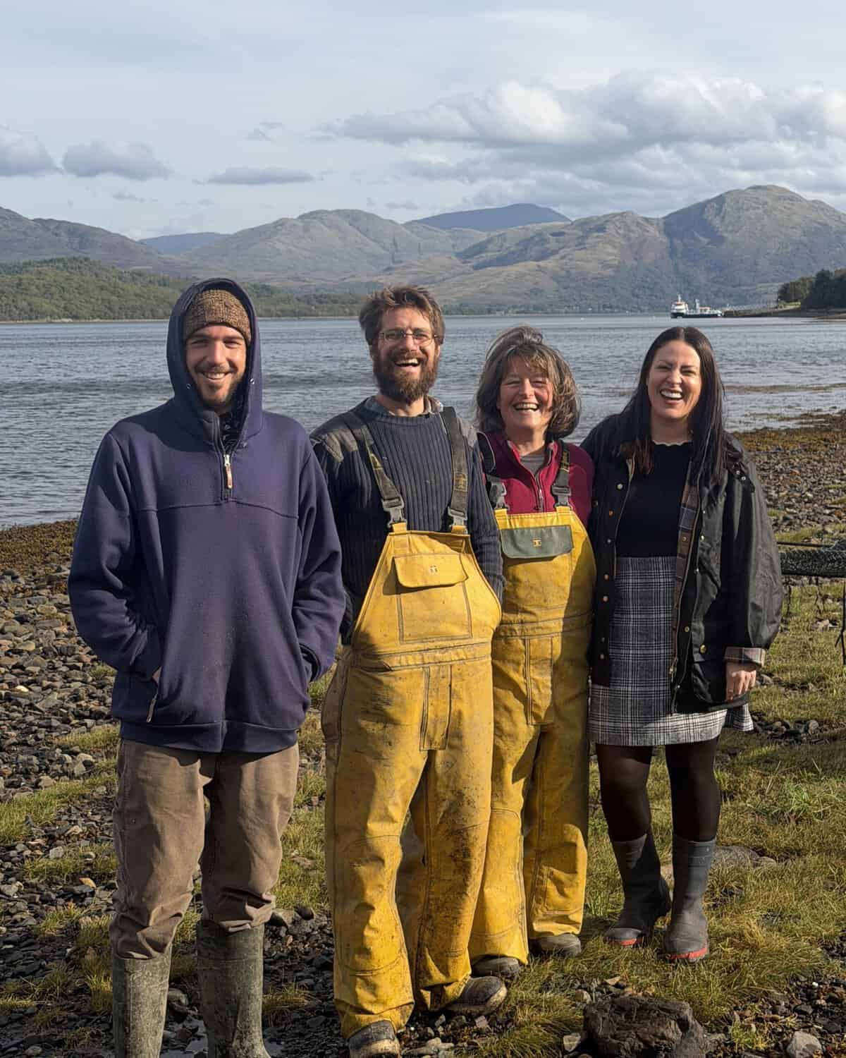 Four people stand on a rocky shore in front of a Scottish loch and mountain range.