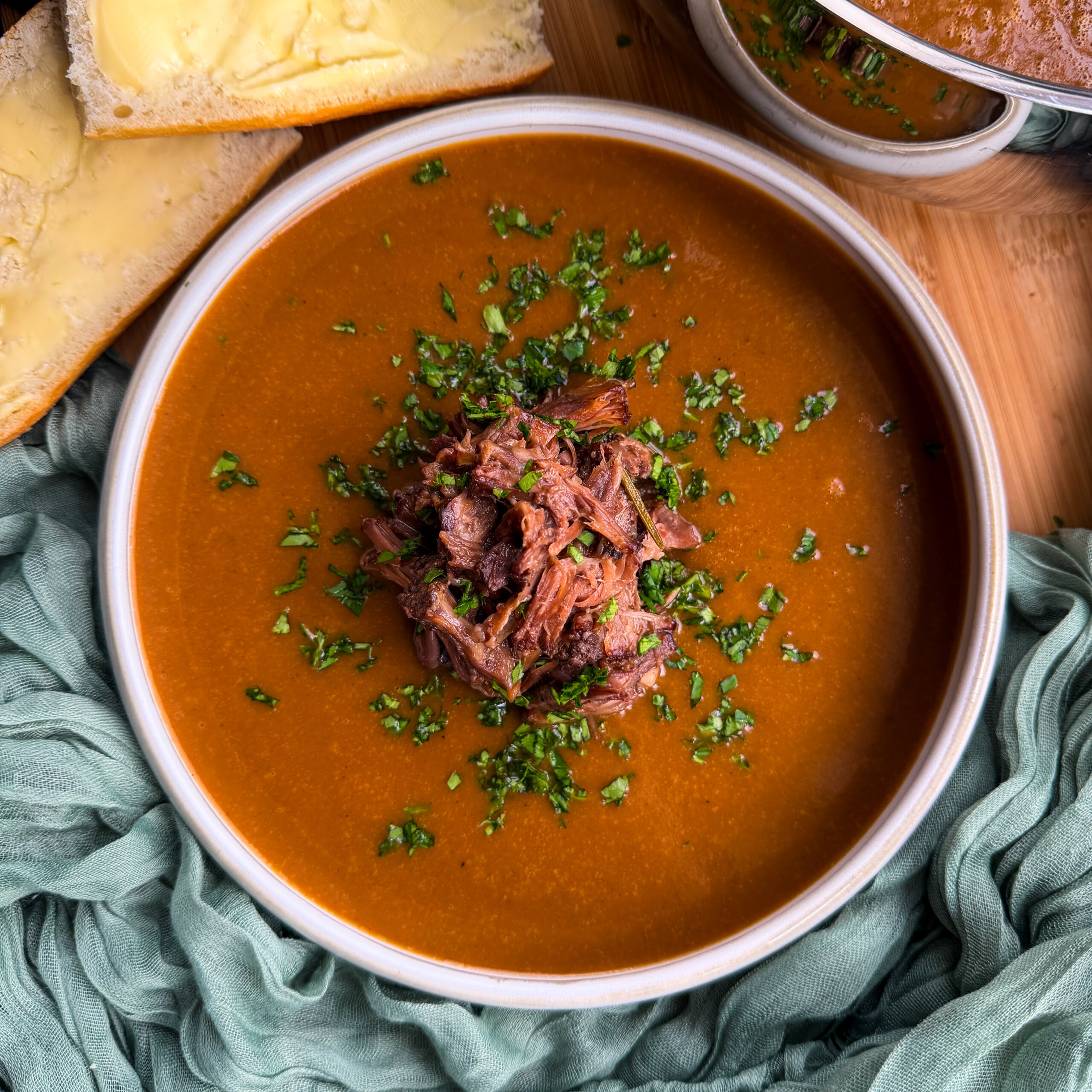 A bowl of oxtail soup topped with shredded beef and herbs, served with slices of buttered bread.