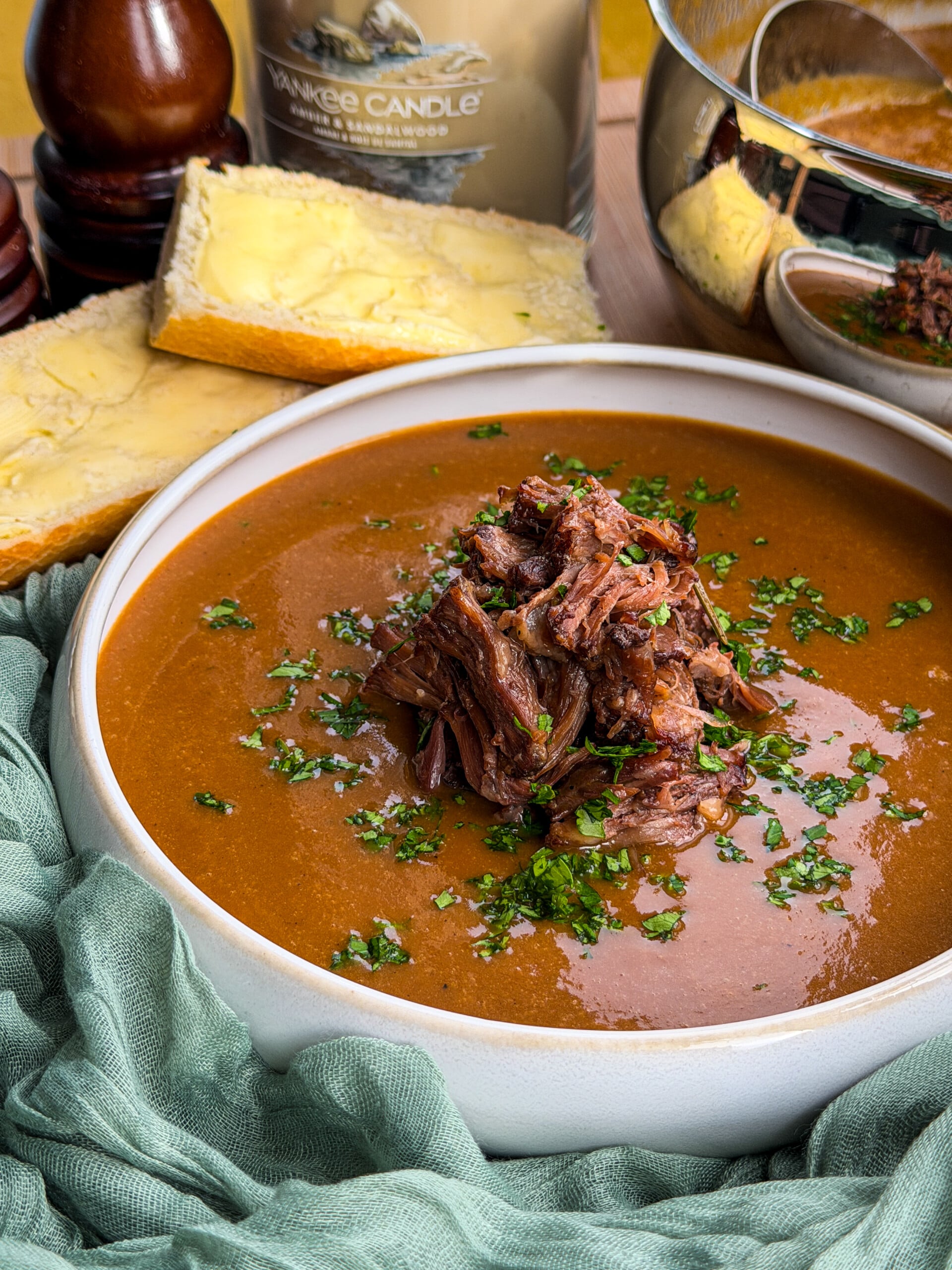 A bowl of oxtail soup topped with shredded meat and chopped herbs, served with slices of buttered bread.
