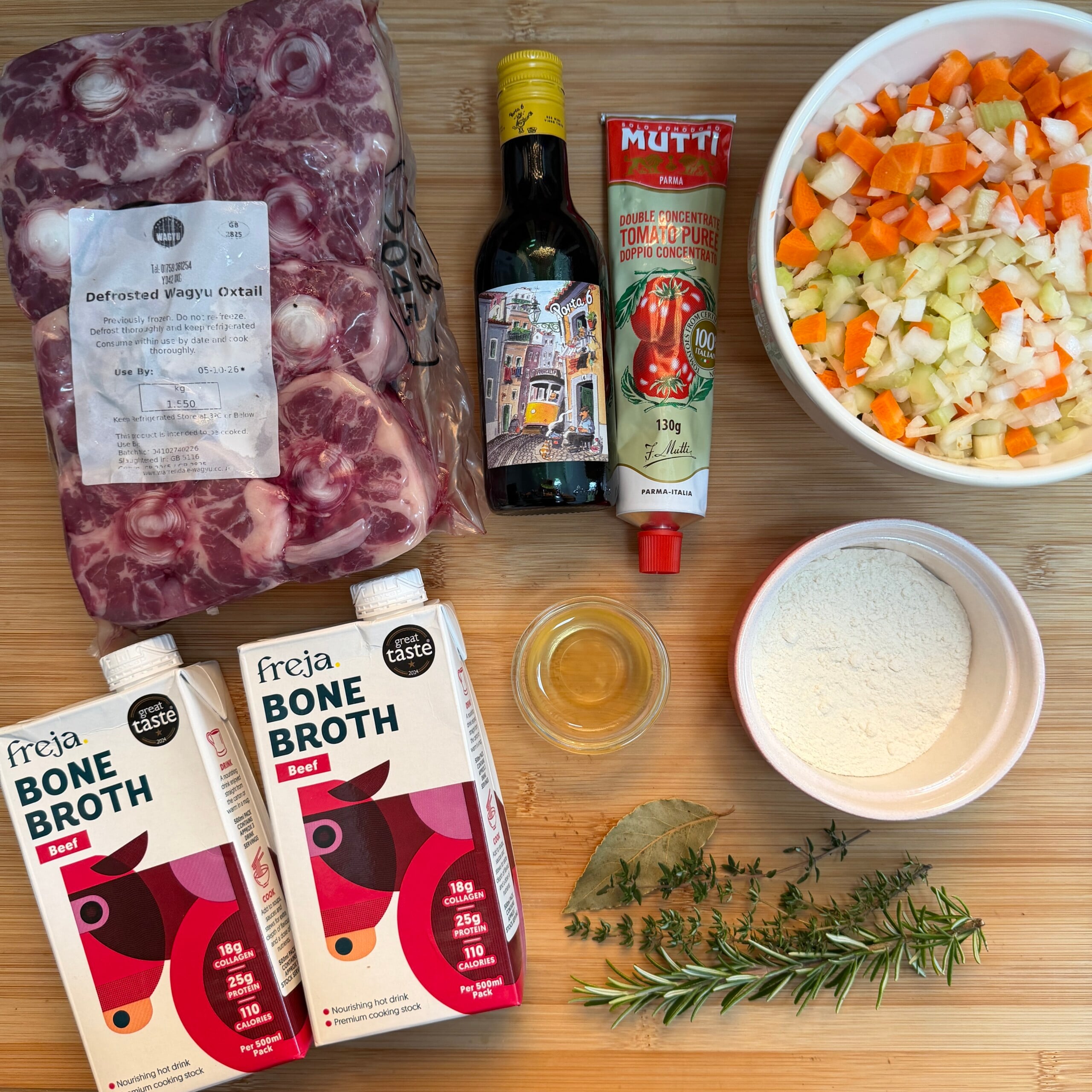 Ingredients for cooking oxtail soup: vacuum-packed oxtail, two cartons of beef stock, a bottle of red wine, a tube of tomato purée, a bowl of diced mirepoix, a bowl of flour, a small dish of oil, and fresh sprigs of rosemary, thyme, and a bay leaf.