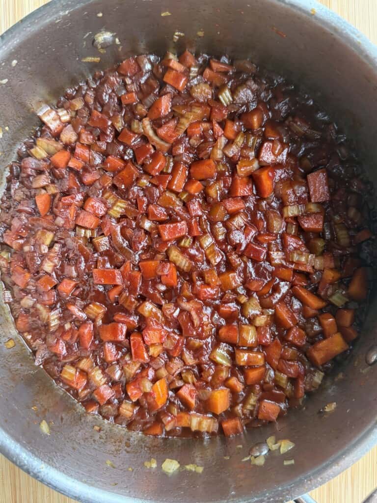A metal saucepan filled with a cooked mixture of diced carrots, celery, and onions in a dark red sauce.