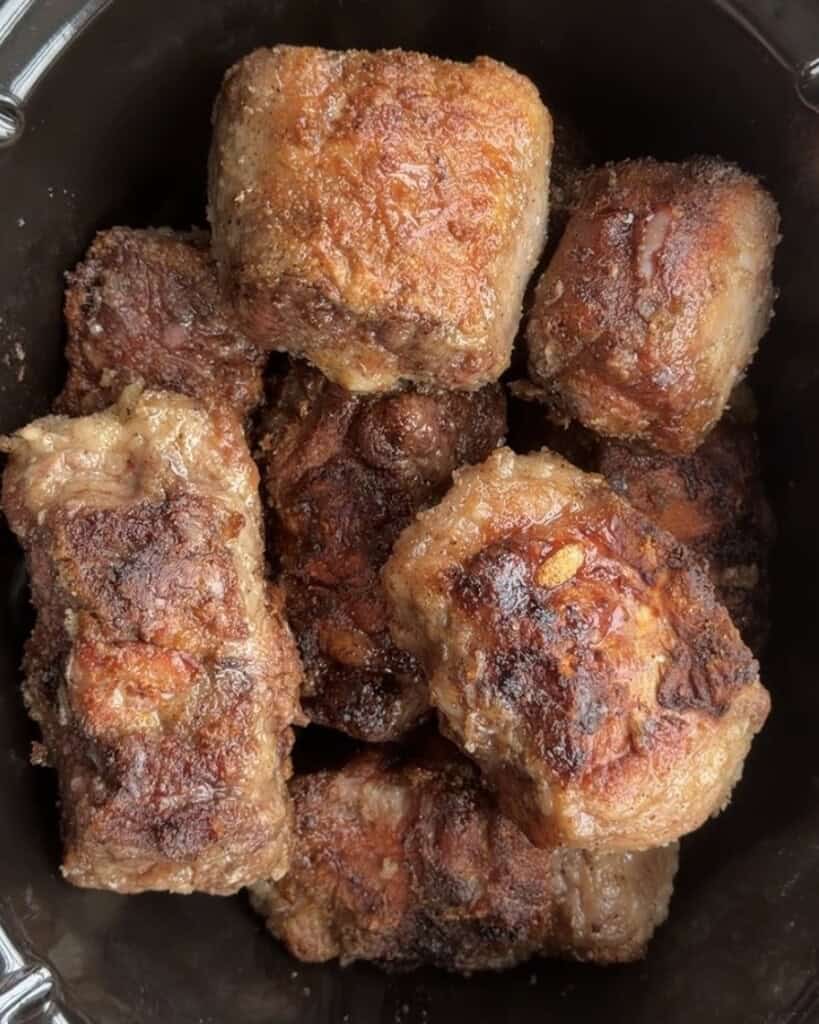 A pile of browned, seared pieces of oxtail in a slow cooker.