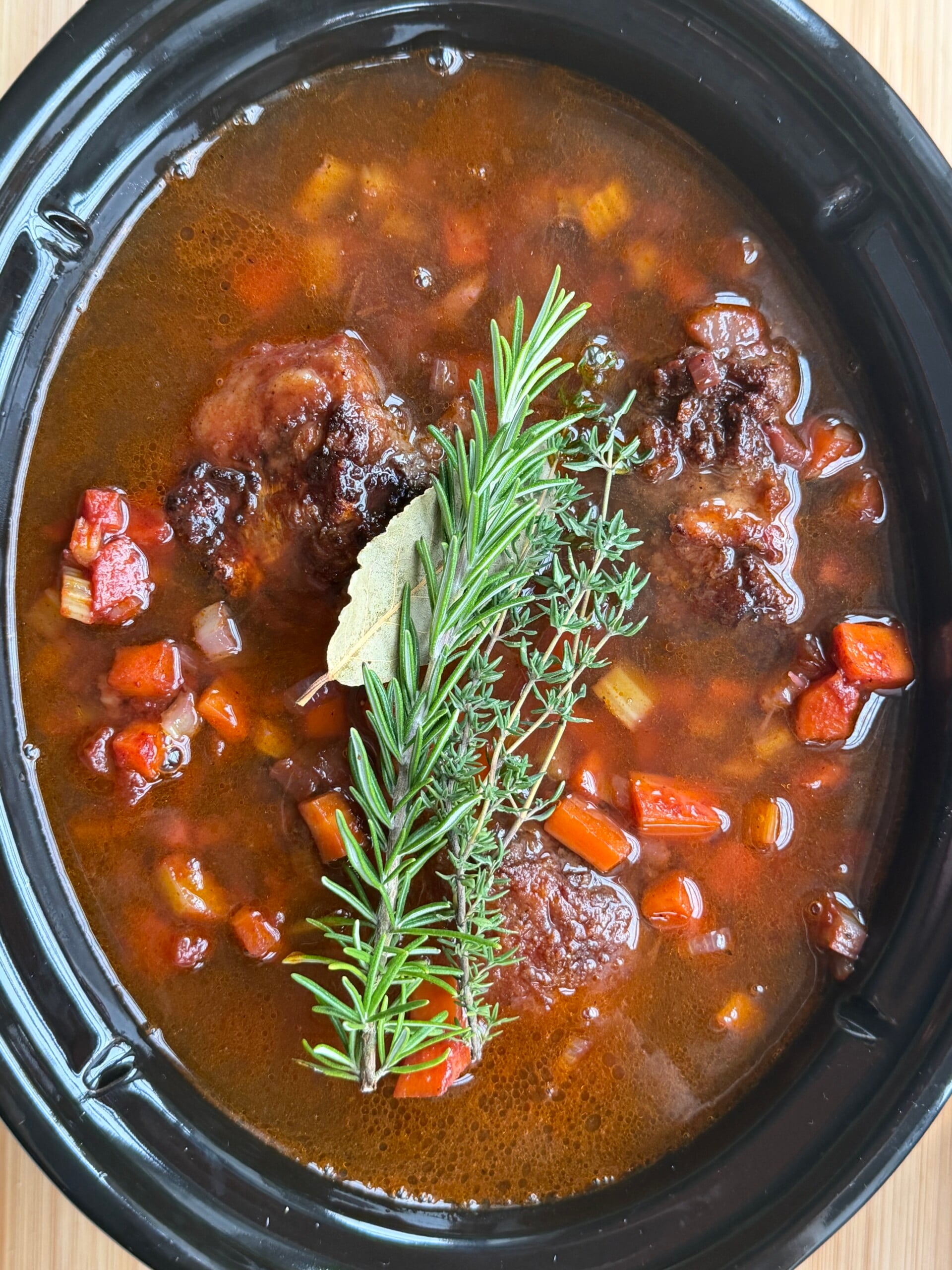 Slow cooker filled with braised meat and diced vegetables in a dark broth, garnished with a sprig of rosemary and a bay leaf.