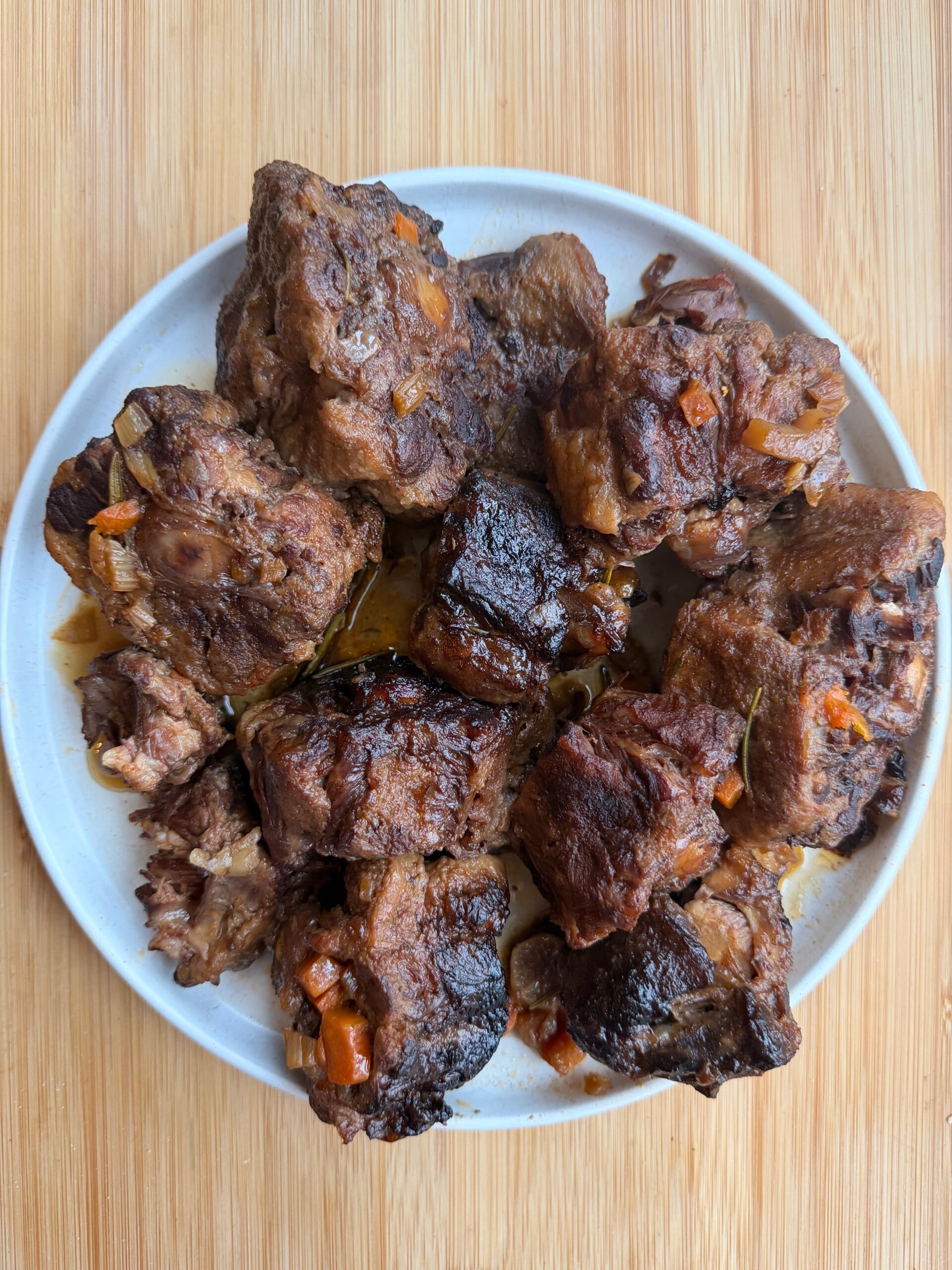 A plate of braised oxtail pieces with bits of carrot and onion on a wooden surface.