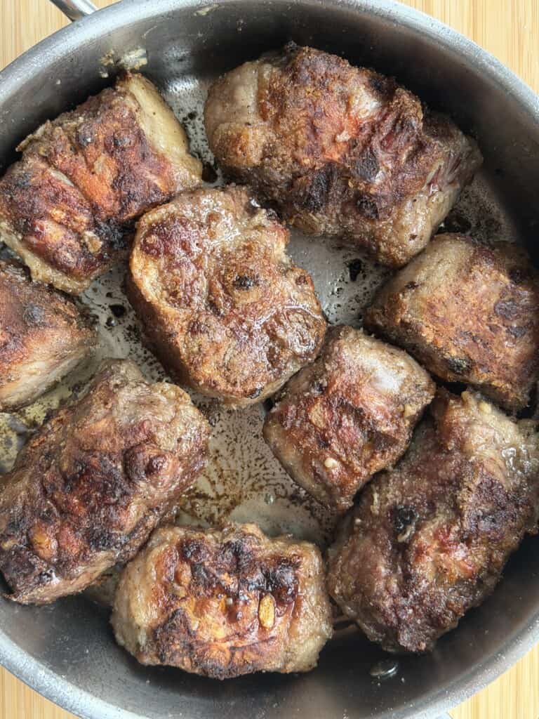 Several pieces of seared oxtail browning in a metal frying pan.