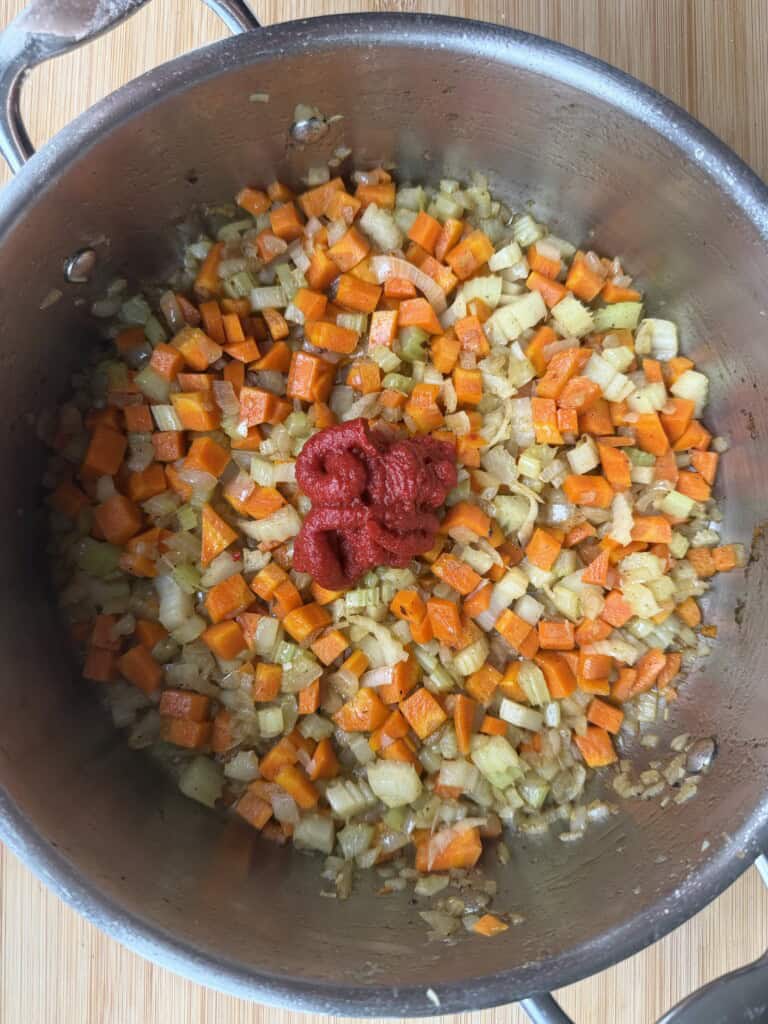 A dollop of tomato purée sits in the centre of a stainless steel saucepan filled with sautéed diced carrots, celery, and onions.