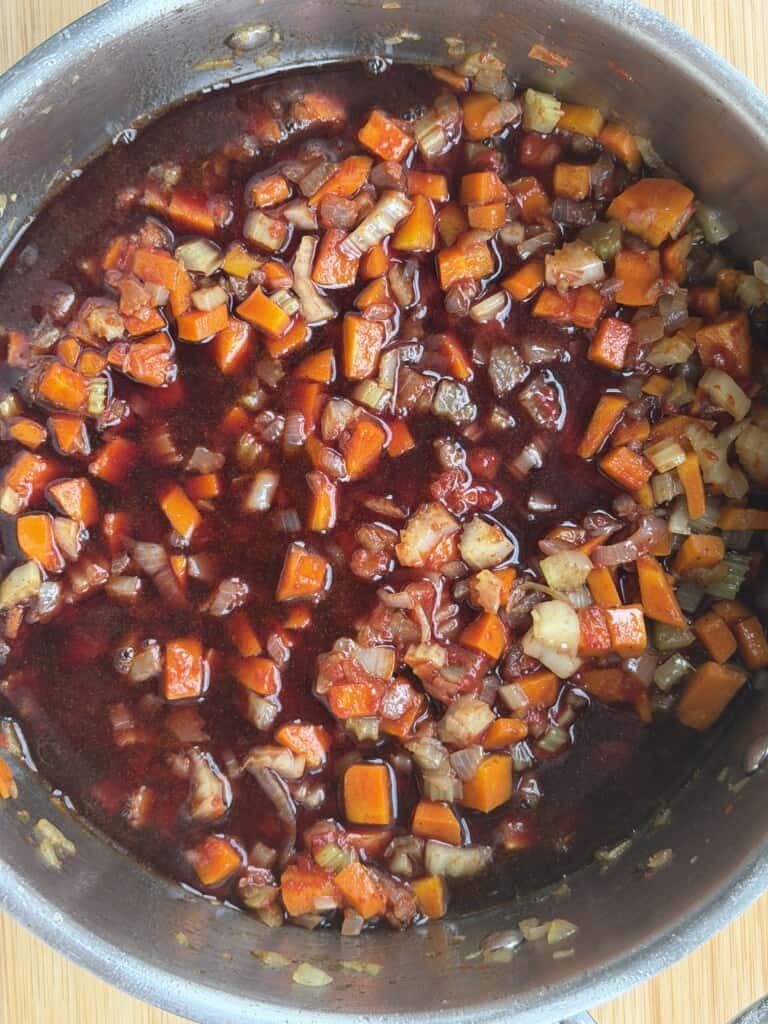 A metal saucepan filled with a dark red, liquid sauce containing diced carrots, onions, and celery.