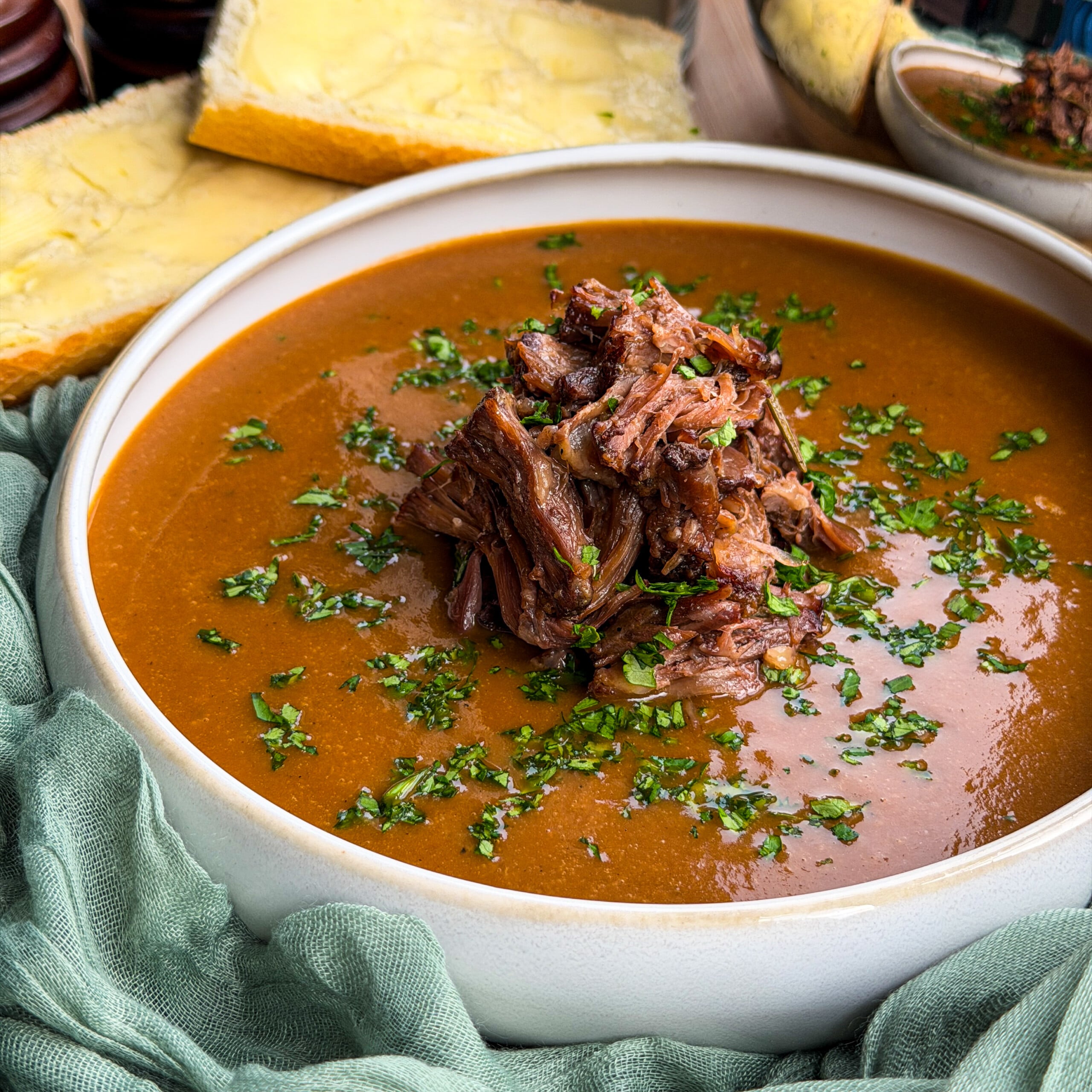 A bowl of oxtail soup topped with shredded beef and herbs, served with slices of buttered bread.