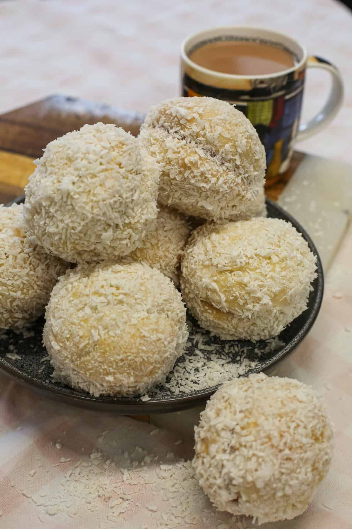 A plate piled with Scottish snowball biscuits coated in desiccated coconut, with a mug of coffee in the background.