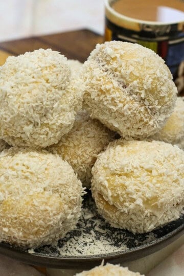 A plate piled with coconut-covered Scottish snowballs next to a mug of coffee.