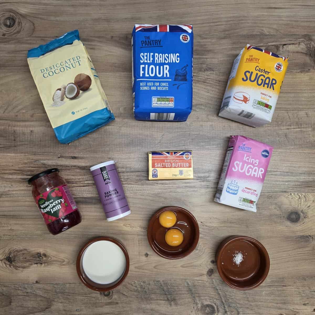 Baking ingredients for scottish snowballs on a wooden surface: bags of desiccated coconut, self-raising flour, caster sugar, and icing sugar; a jar of raspberry jam, a tub of baking powder, a block of butter, two egg yolks, milk, and a pinch of salt.