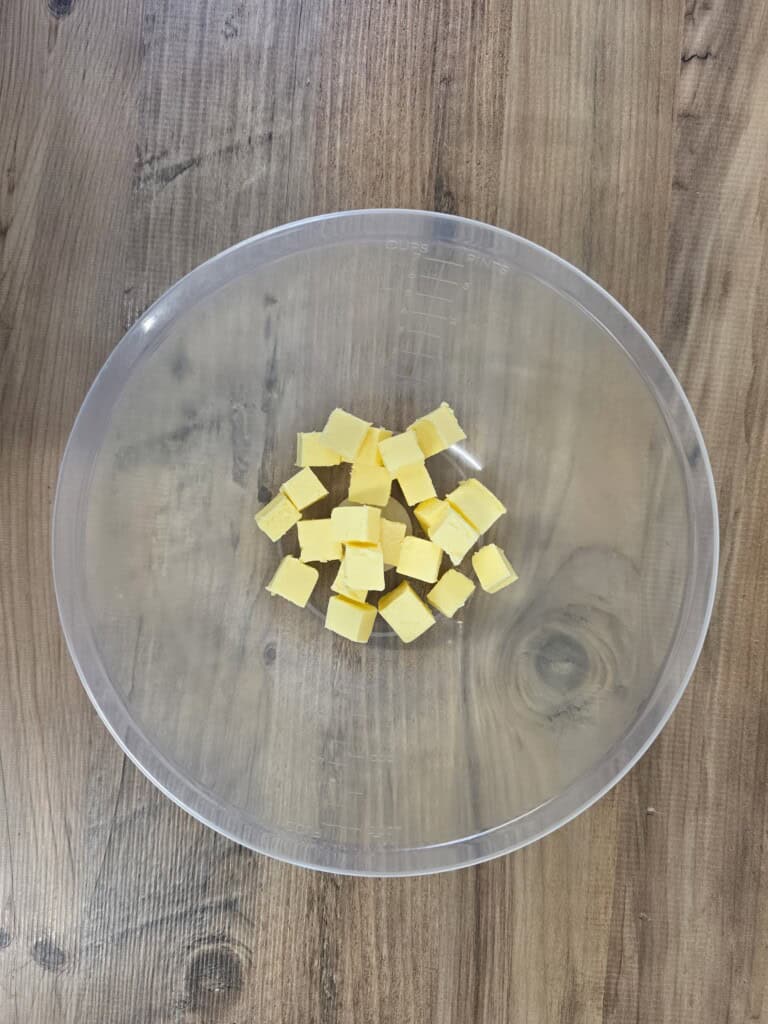 Cubes of butter in a clear plastic mixing bowl on a wooden surface.