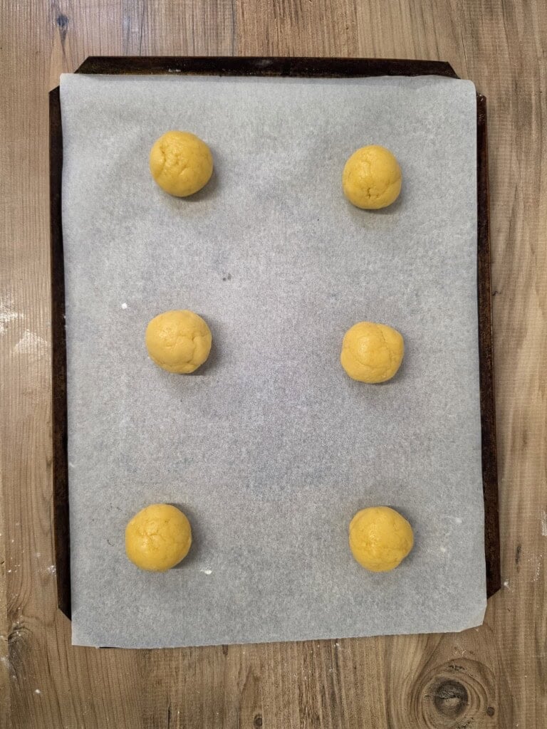 Six balls of raw cookie dough arranged on a baking tray lined with greaseproof paper.