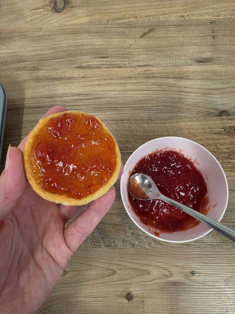 A hand holds a toasted round bread topped with strawberry jam next to a small bowl of jam with a spoon inside.