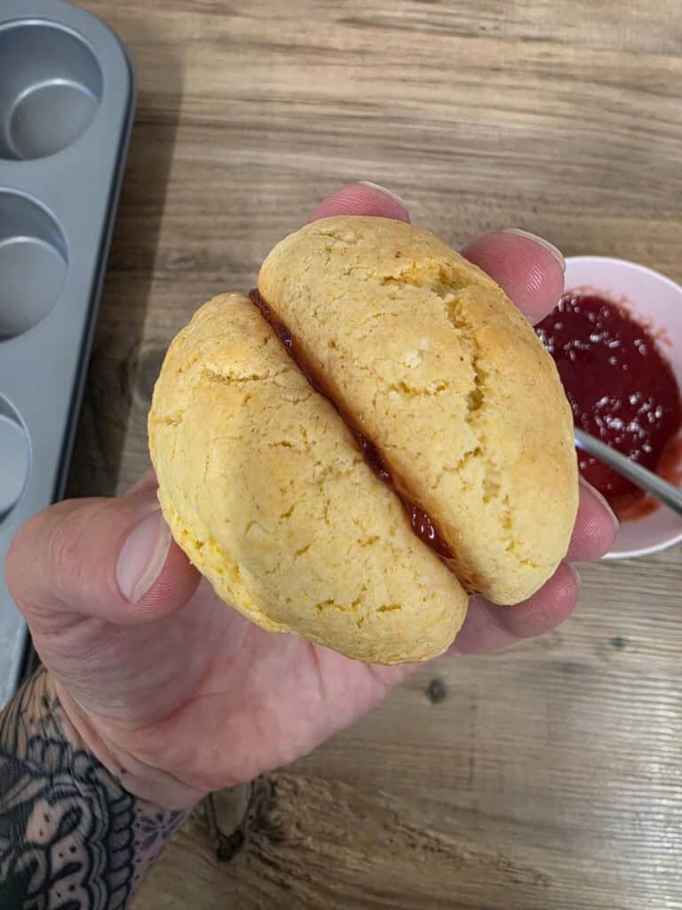 A hand holds a golden, round scone split in half with red jam in the centre.