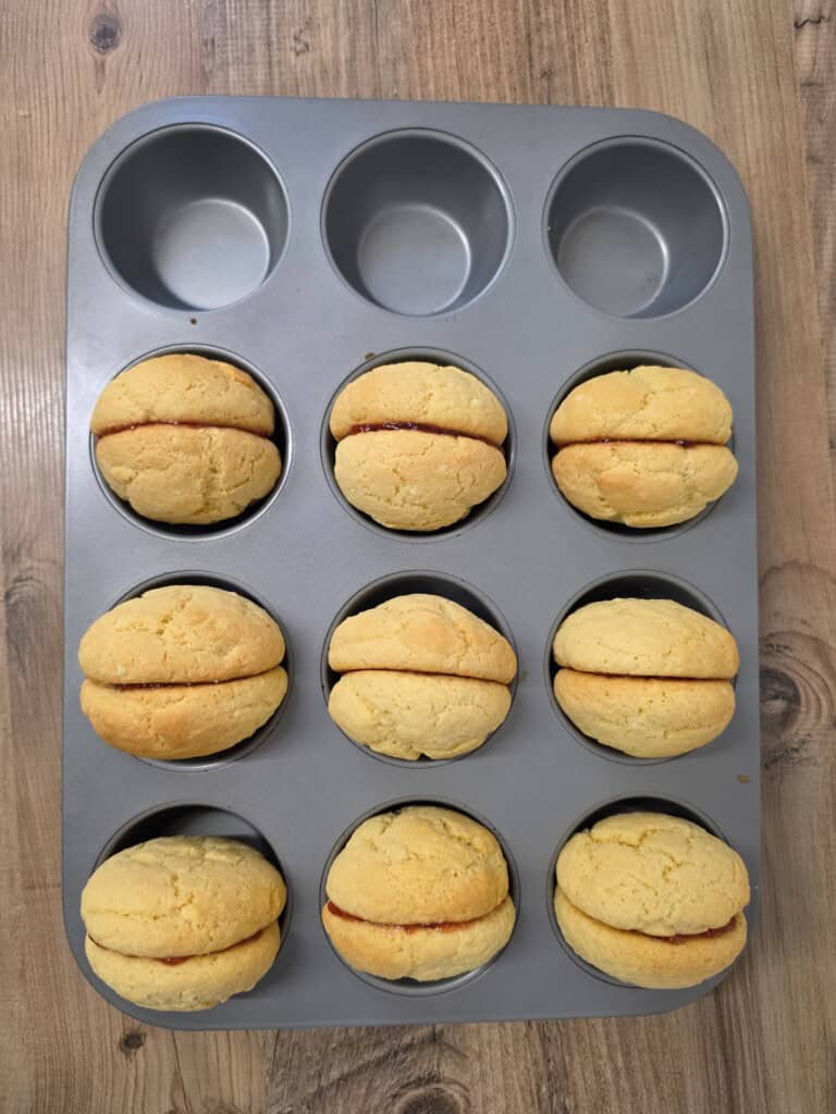 Eight biscuit sandwiches filled with red jam resting in a muffin tin.