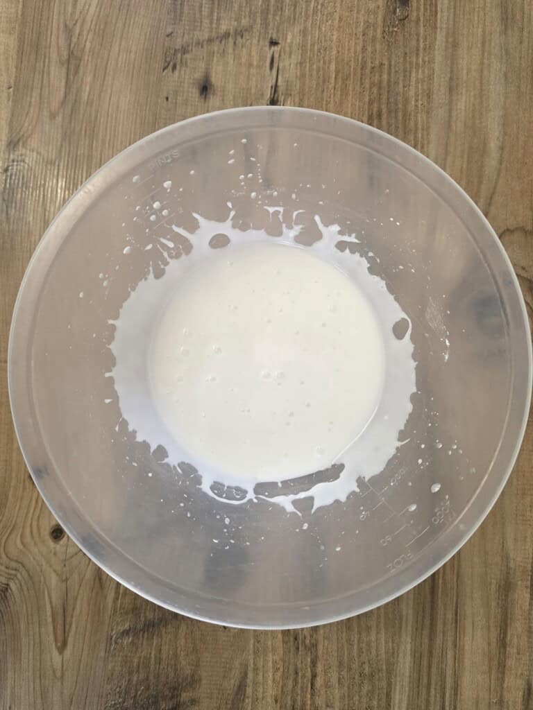 A translucent plastic mixing bowl containing a thin white liquid, with splashes dried onto the inner sides, sitting on a wooden surface.