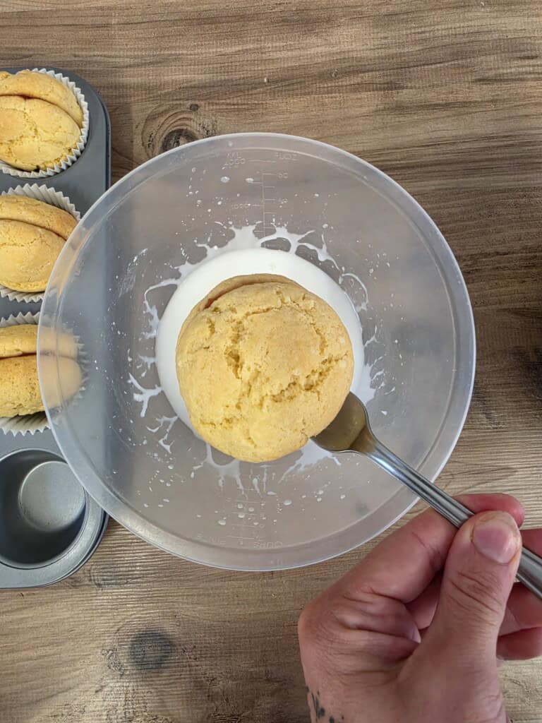 A hand uses a spoon to dip a round, golden biscuit into a bowl of white icing, next to a muffin tin containing similar biscuits.