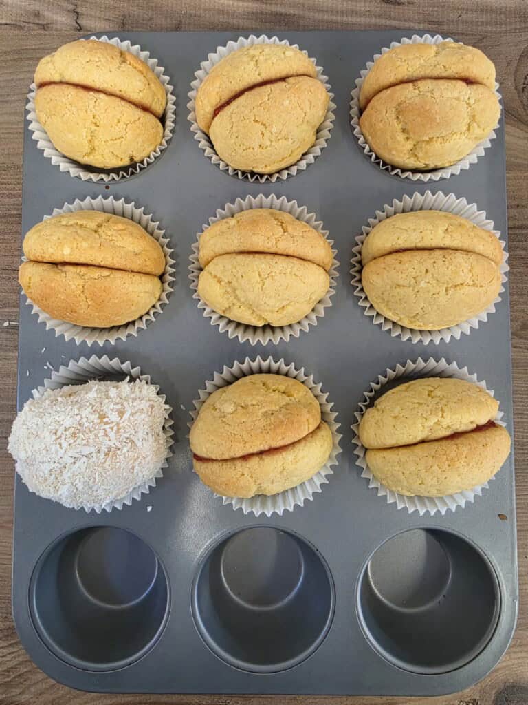 A muffin tray containing eight round sandwich biscuits with jam filling, and one biscuit coated in desiccated coconut.