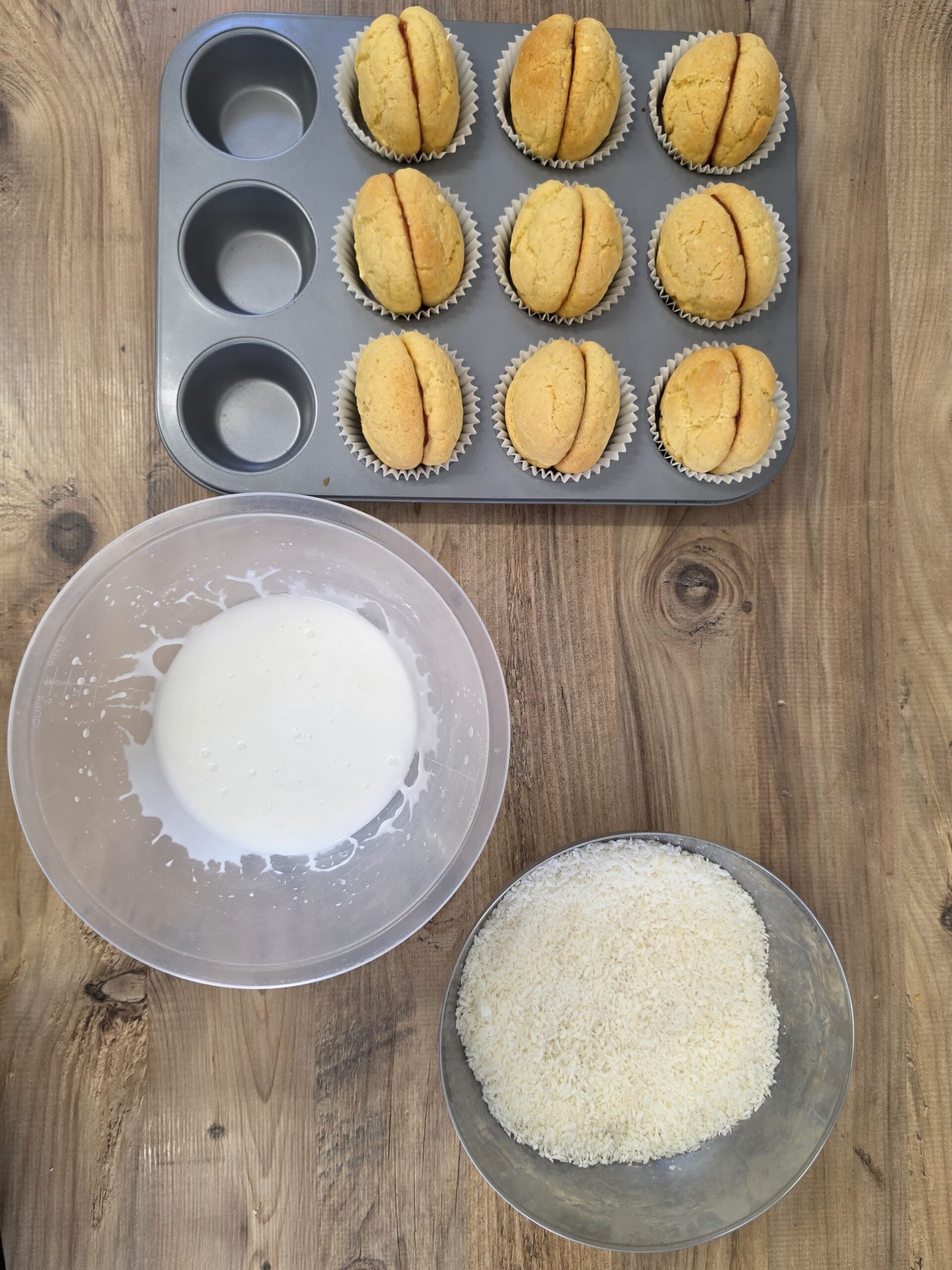 A baking tray filled with nine sandwich biscuits in paper cases, with a bowl of white icing and a bowl of desiccated coconut on a wooden surface.