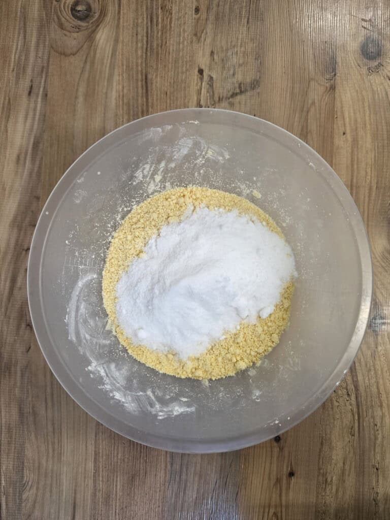A pile of white sugar sitting on top of yellow crumbly dough in a clear mixing bowl.