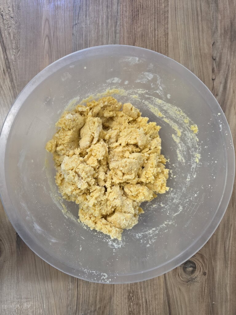A clear mixing bowl containing a crumbly, yellow biscuit or pastry dough.