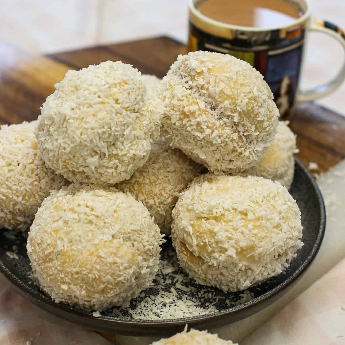 A plate piled with coconut-covered Scottish snowballs next to a mug of coffee.
