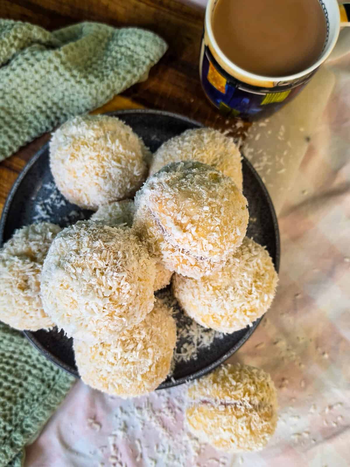 A pile of coconut-covered Scottish snowballs on a dark plate, next to a mug of coffee.
